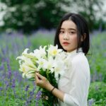Side view of charming ethnic female with long dark hair standing with bunch of Easter lilies in blooming field and looking at camera