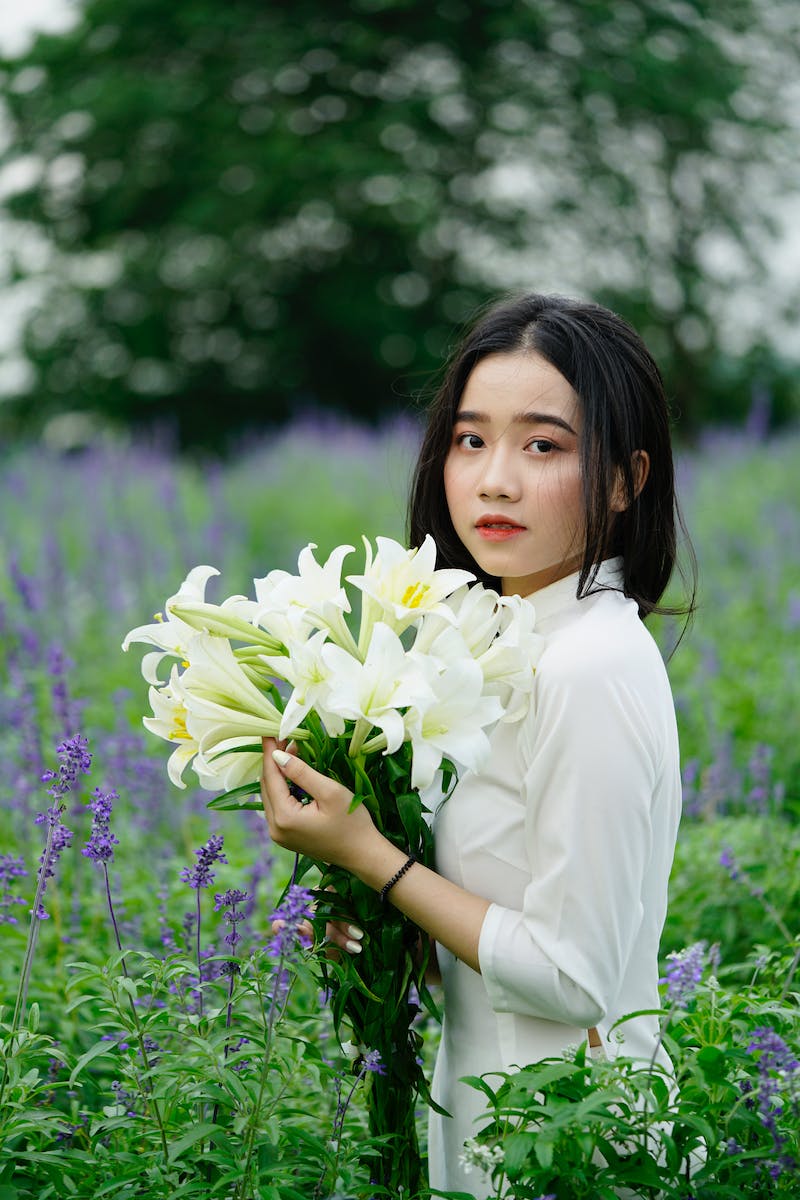 Side view of charming ethnic female with long dark hair standing with bunch of Easter lilies in blooming field and looking at camera